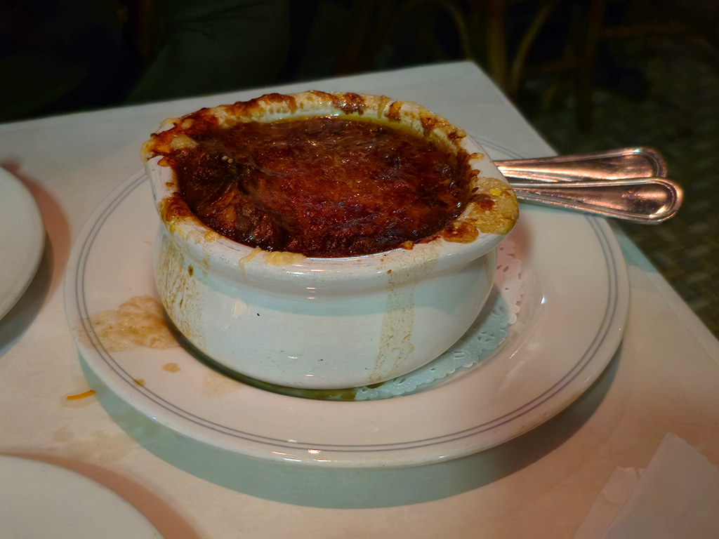 A photograph of a small bowl of onion soup with a dry, crusted layer sitting on top of the soup, many globs and streaks of soup that had overflowed around the edges of the bowl and dripped down the sides, and drops of dried soup on the saucer under the bowl.